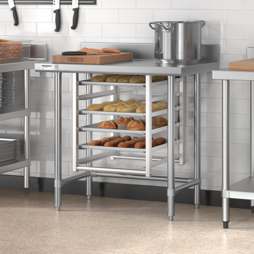 A stainless steel work table with an open base, featuring a 4-inch backsplash, multiple trays of baked goods stored underneath, and kitchen items on top.