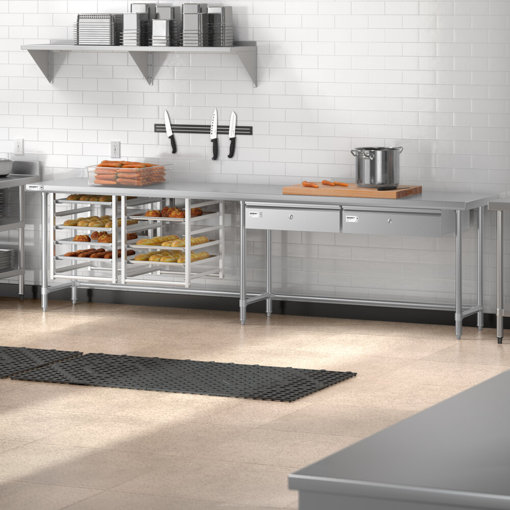 A stainless steel commercial kitchen work table with an open base, drawers, and shelves holding trays of food, set against a white tiled wall with knives, a pot, and cutting boards on the countertop.