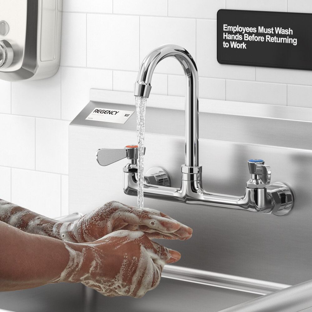 A person is washing their soapy hands under a wall-mounted, chrome faucet with a gooseneck spout in a commercial sink area.