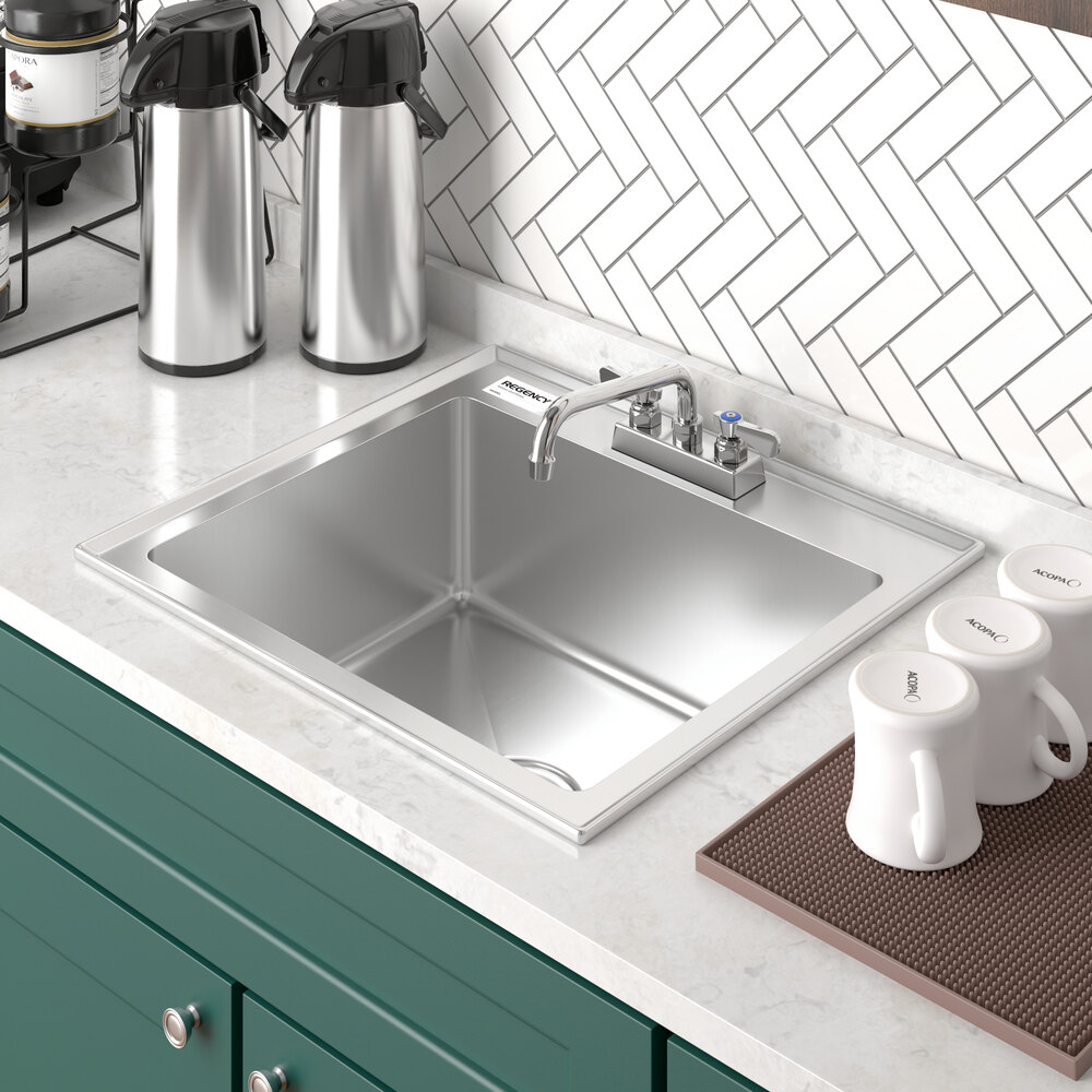 A stainless steel single-basin sink with a faucet, set in a white countertop with green cabinetry and surrounded by coffee mugs and dispensers.