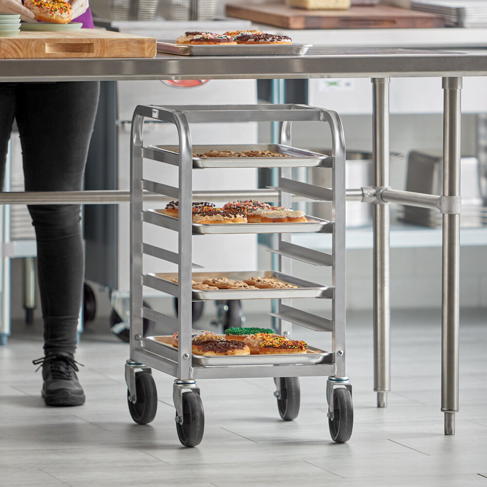 A metal rack on wheels holding several trays of baked goods, positioned under a stainless steel table in a kitchen setting.