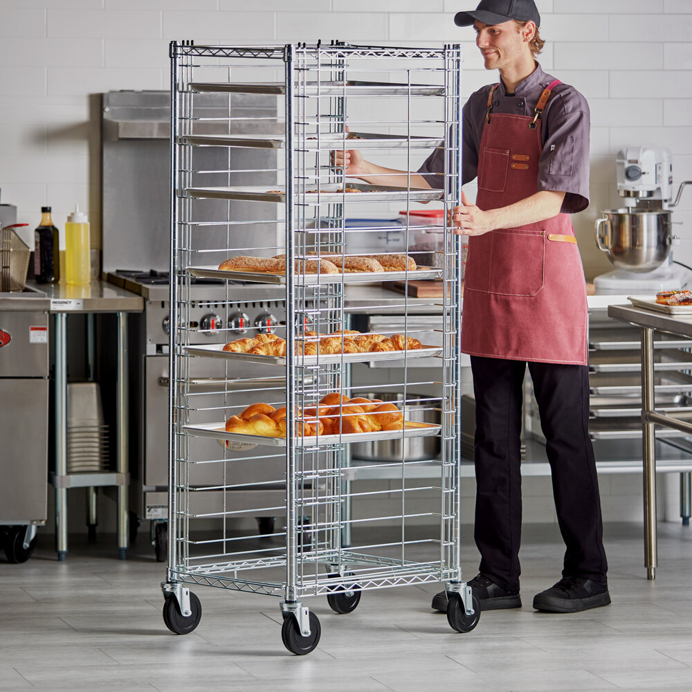 A person in a commercial kitchen is pushing a tall, metal wire rack on wheels that holds several trays of baked goods.