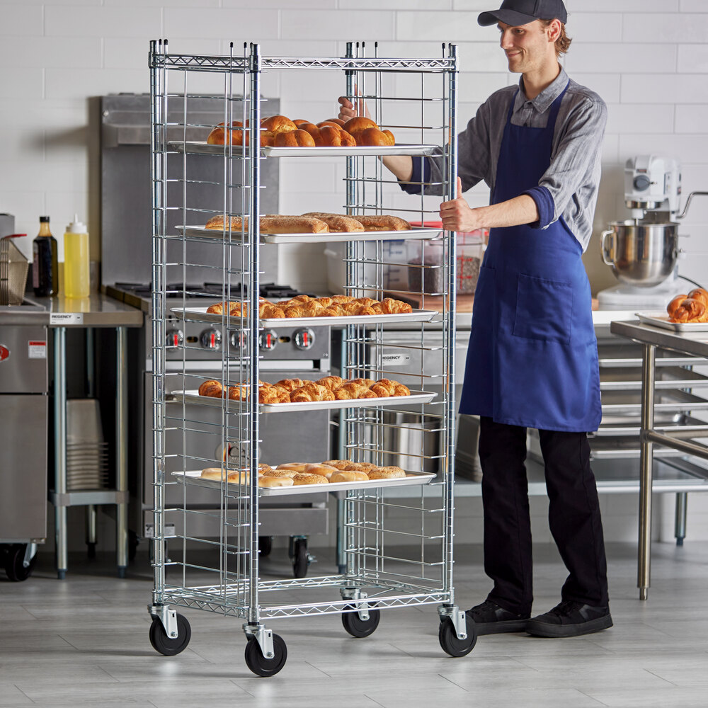 A person in a commercial kitchen is pushing a tall metal rack on wheels, which holds multiple trays of baked goods such as rolls and croissants.