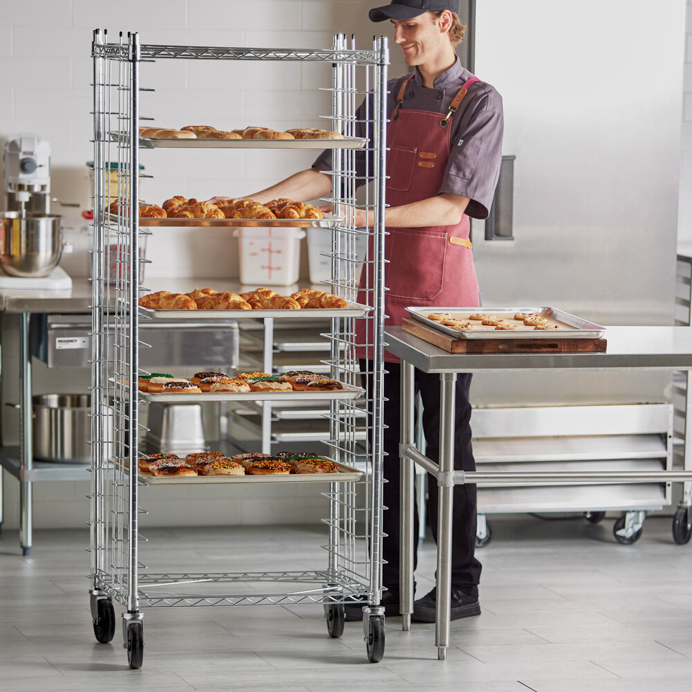 A person in a commercial kitchen stands next to a tall, metal wire rack on wheels, which holds multiple trays of baked goods such as pastries and donuts.