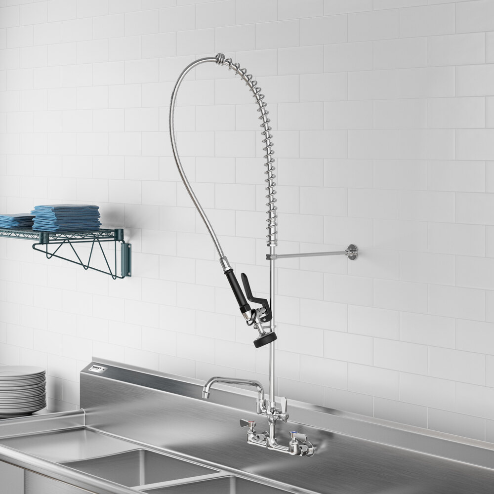 A commercial-style wall-mounted pre-rinse faucet with a flexible hose and spray nozzle, installed above a stainless steel sink in a kitchen setting.