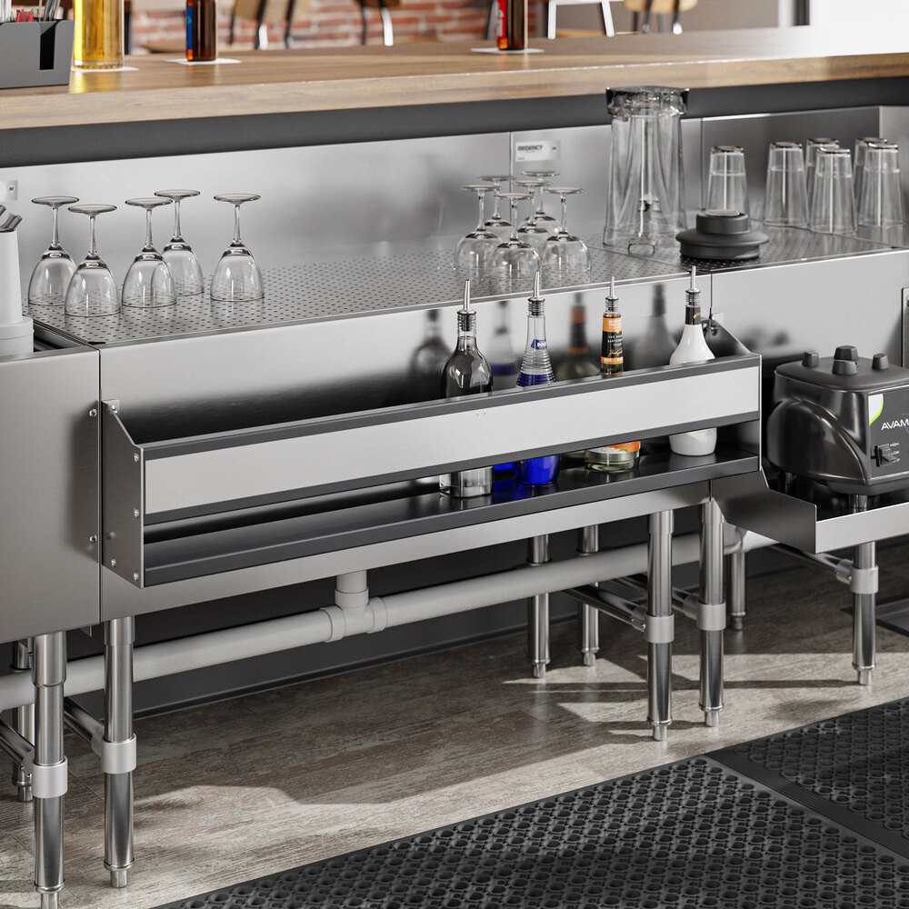A stainless steel bar setup with a single tier speed rail holding bottles, glassware on a perforated shelf, and visible plumbing underneath.