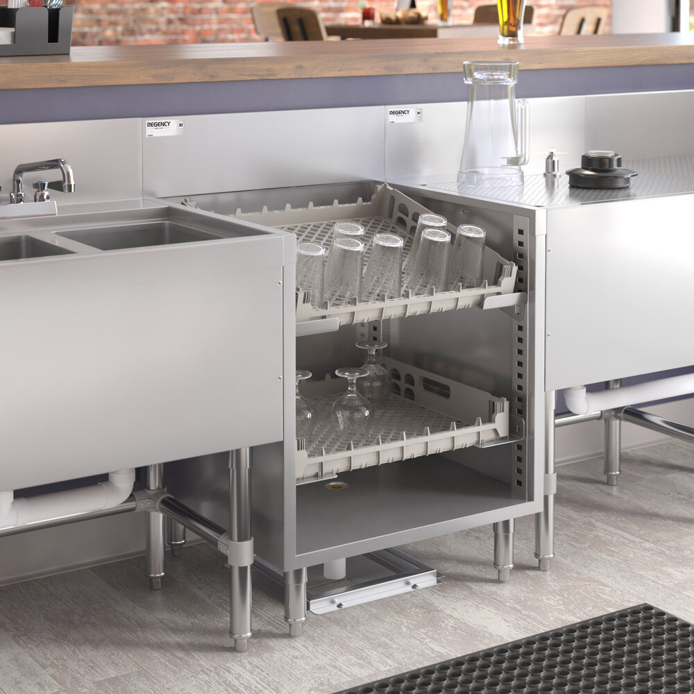 A stainless steel open storage unit with two pull-out racks holding glassware, situated between a sink and a countertop in a commercial kitchen setting.