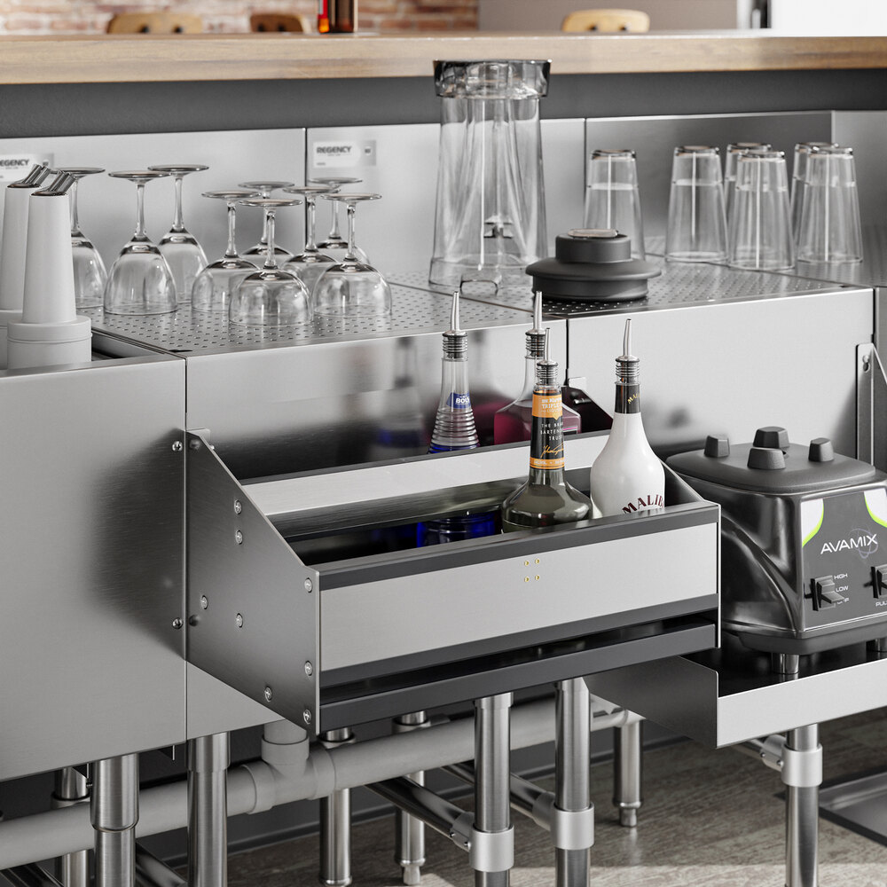 A stainless steel double-tier speed rail holding several bottles, situated beneath a bar counter with glassware and a blender nearby.