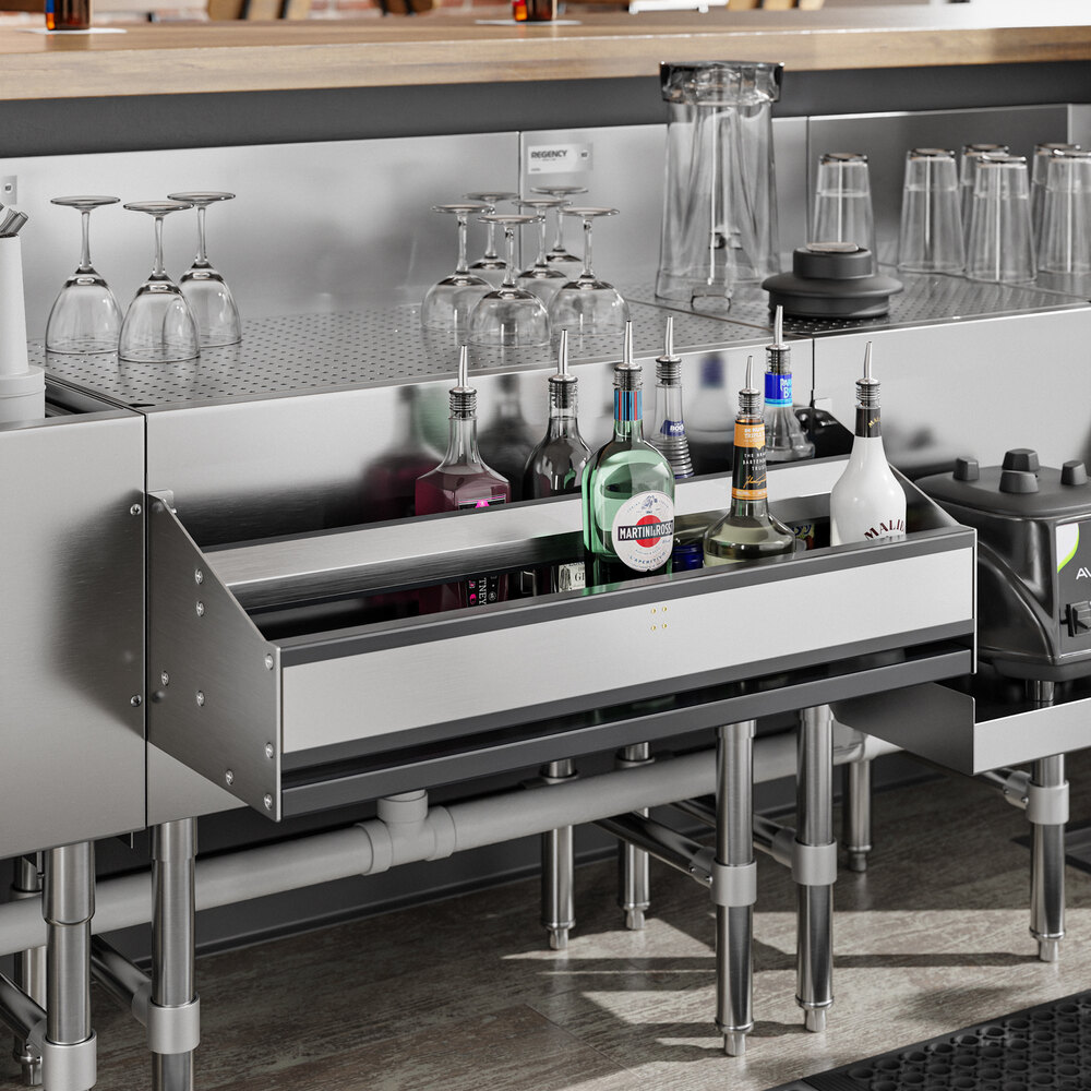 A stainless steel double-tier speed rail filled with various liquor bottles, installed under a bar counter with glassware and pitchers on top.