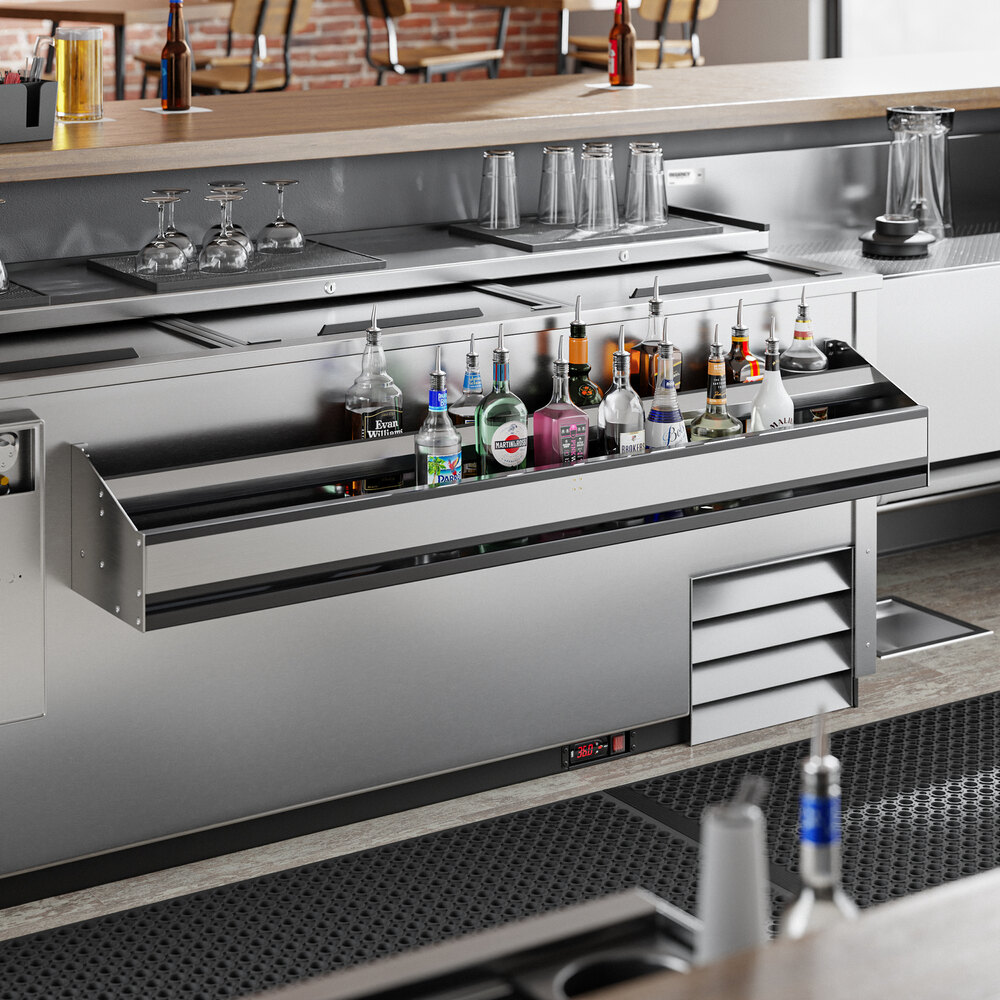 A stainless steel double-tier speed rail filled with various liquor bottles is mounted below a bar counter, with glassware and other bar equipment visible nearby.