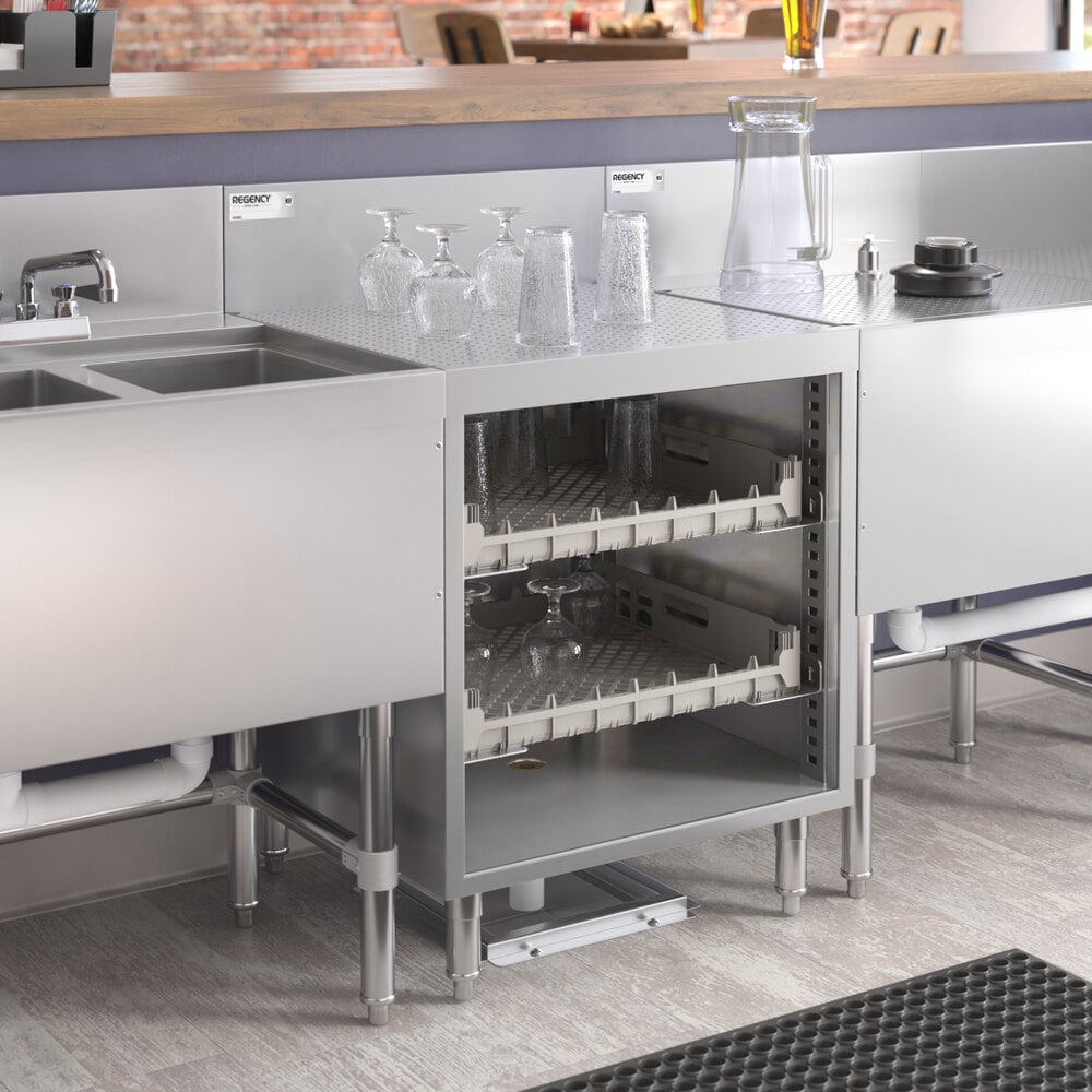 A stainless steel storage unit with two shelves holding glassware, situated between a sink and a countertop in a commercial kitchen or bar setting.