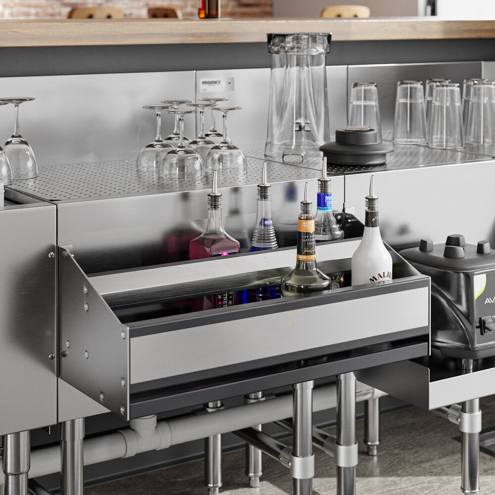 A stainless steel double-tier speed rail is mounted under a bar counter, holding various liquor bottles, with glassware and pitchers arranged on the counter above.