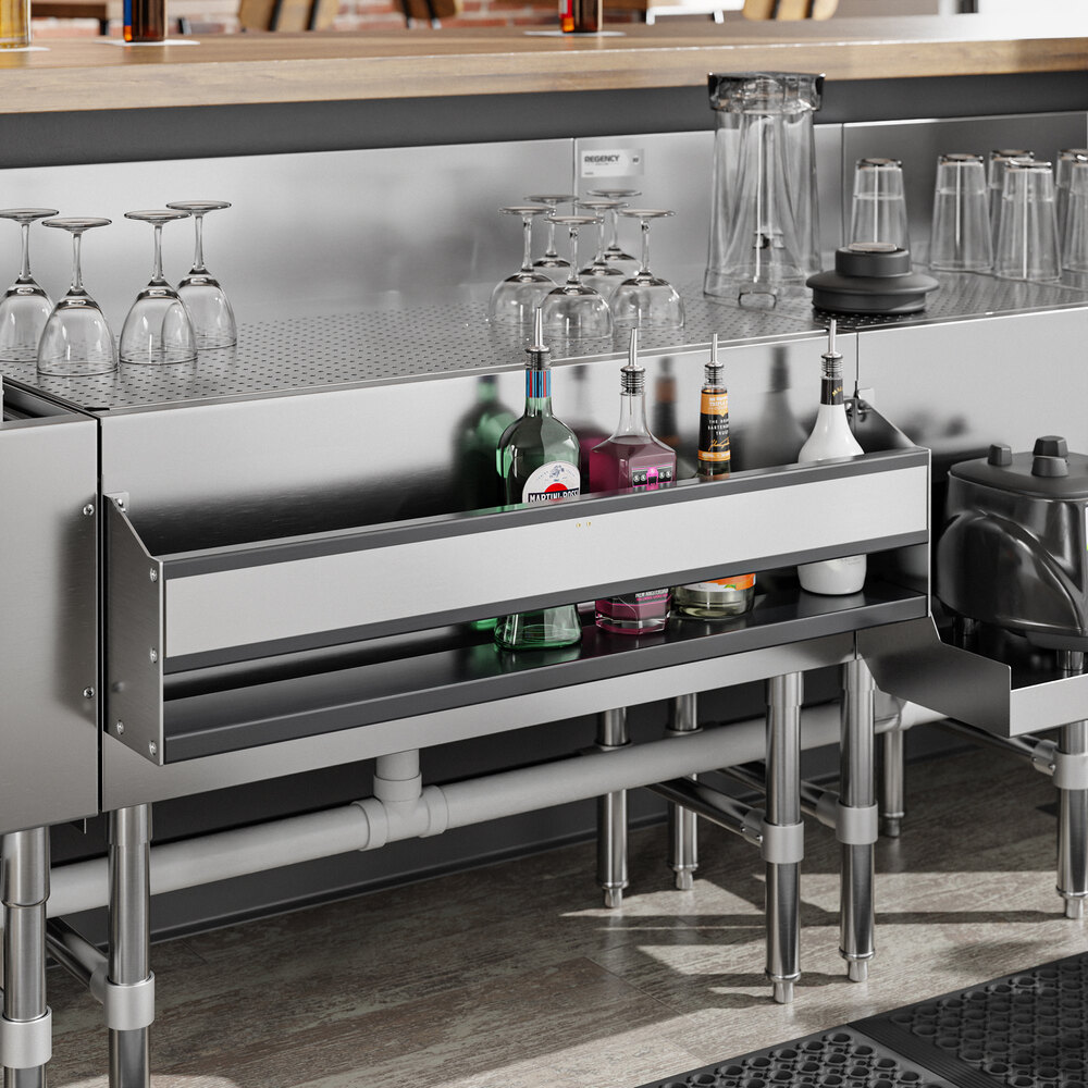 A stainless steel single-tier speed rail attached to a bar counter, holding several bottles and bar supplies, with glassware stored above.