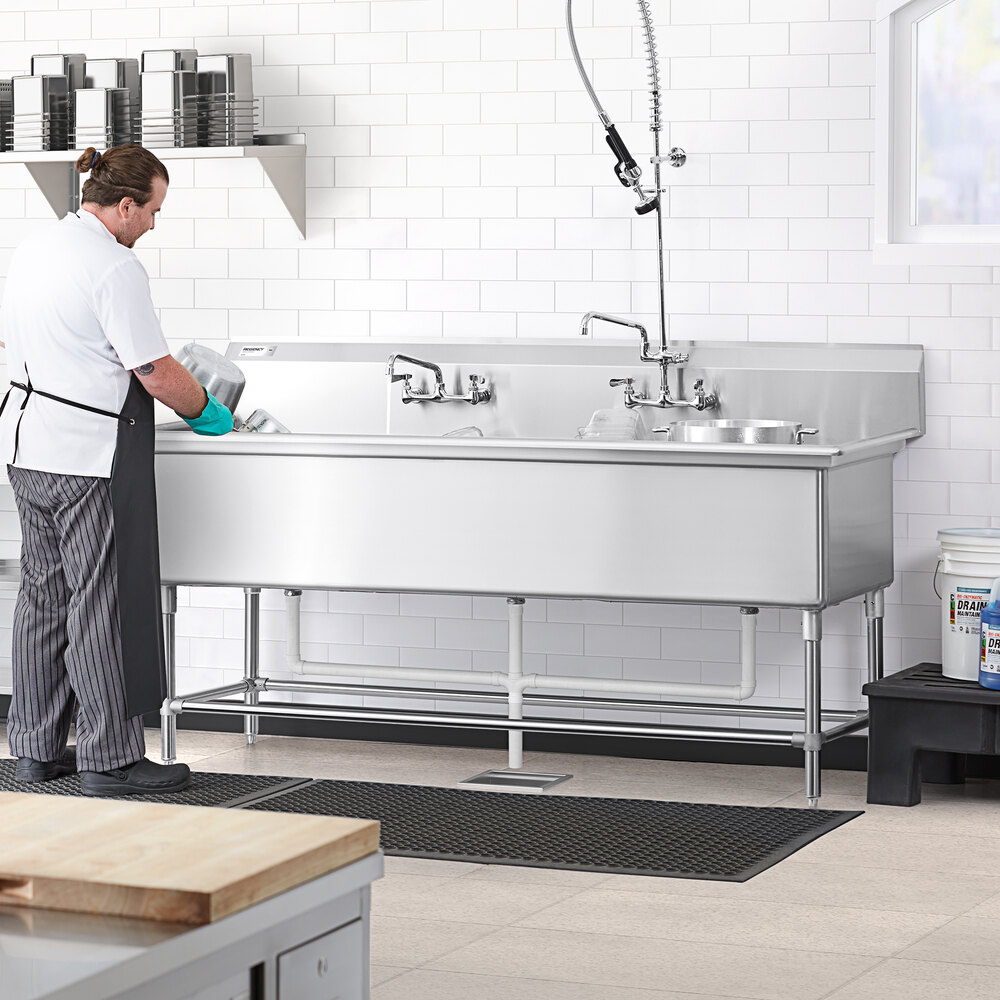 A person is washing dishes at a large, stainless steel, three-compartment sink in a commercial kitchen setting.