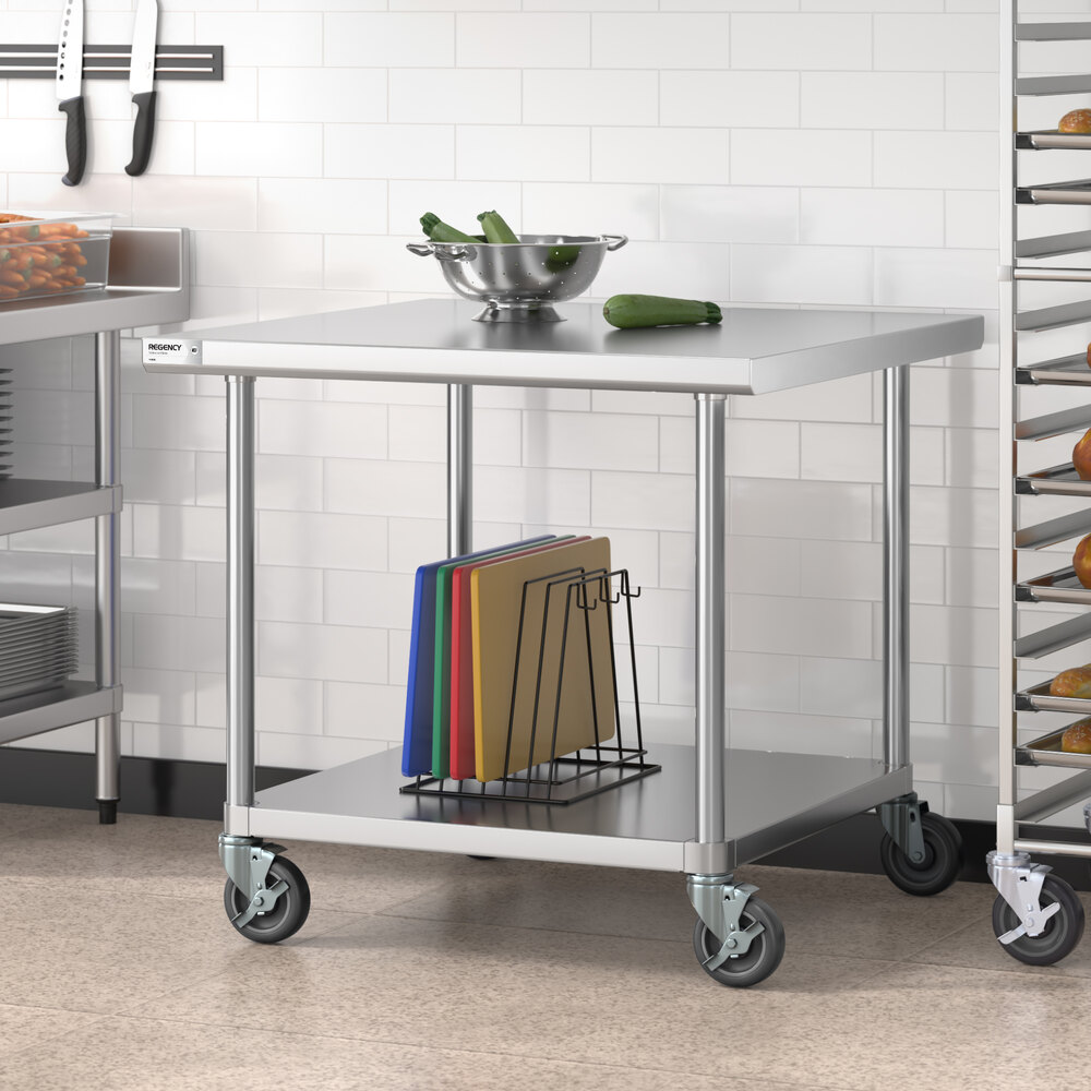 A stainless steel work table with an undershelf, casters, a metal bowl with vegetables on top, and cutting boards stored below.
