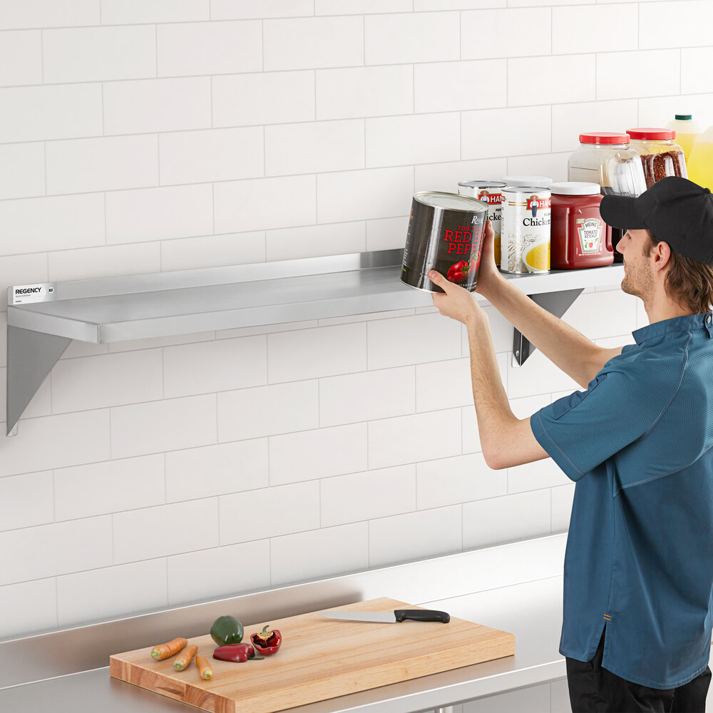 A person is placing a large can on a long, wall-mounted stainless steel shelf in a kitchen setting, with various food containers and jars also on the shelf.