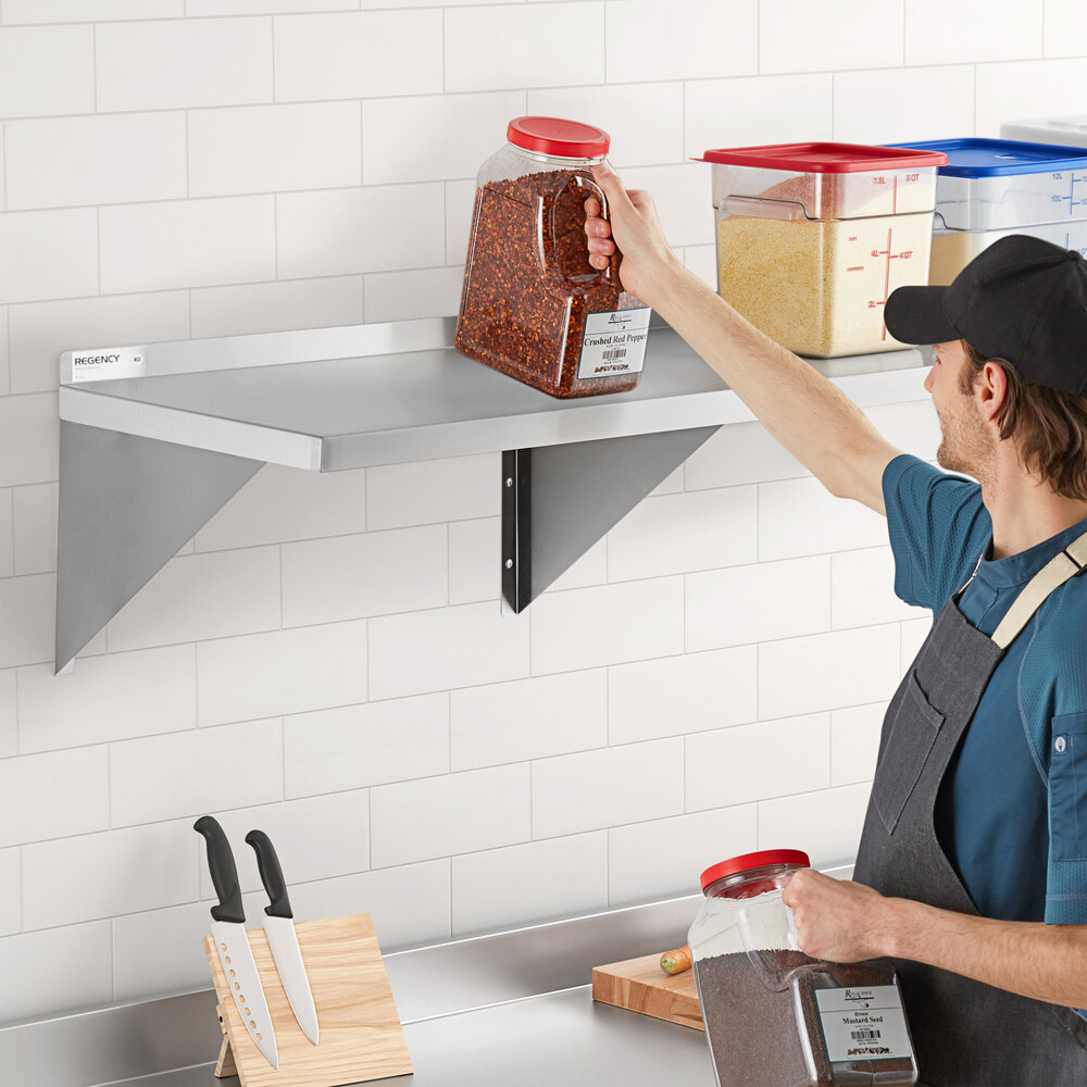 A person is placing a large container of spices on a stainless steel wall shelf mounted on a tiled kitchen wall, with other containers and kitchen knives visible nearby.