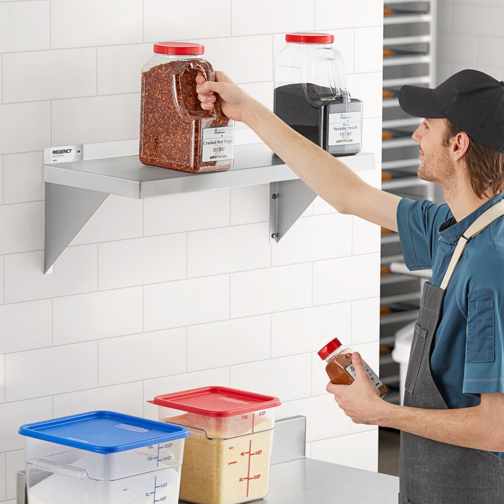A person is reaching for a large container of spices on a stainless steel wall shelf mounted on a white tiled wall.