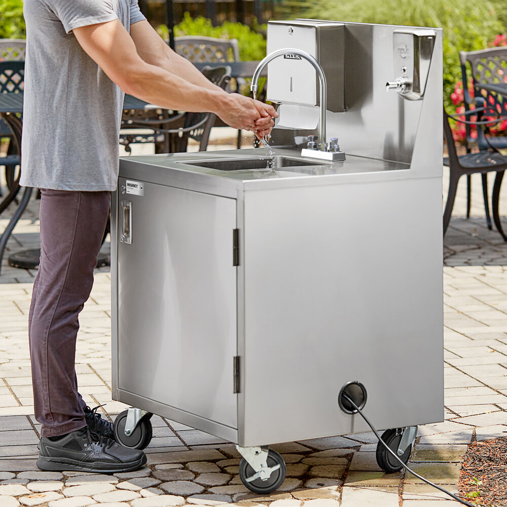 A person is washing their hands at a stainless steel portable hand sink cart with a gooseneck faucet, paper towel dispenser, and wheels for mobility.