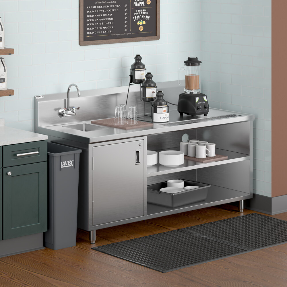 A stainless steel beverage station with a left-side sink, gooseneck faucet, open shelves holding cups and dishes, a blender, and condiment dispensers, set against a light blue tiled wall.