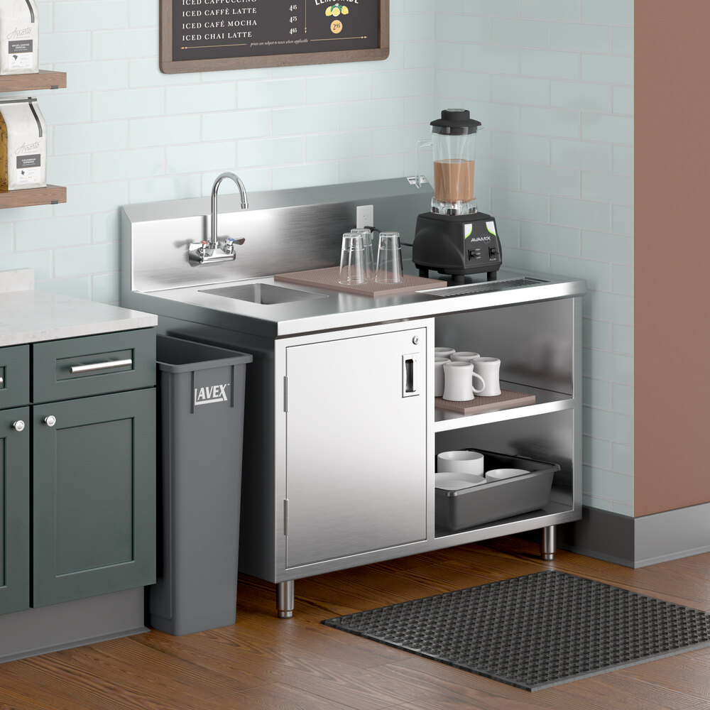 A stainless steel beverage table with a left-side sink, gooseneck faucet, open shelves, and a blender on top, situated in a small kitchen or café area.