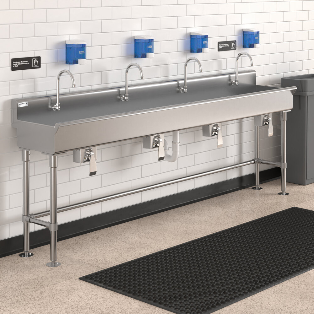 A long stainless steel hand sink with four faucets, mounted against a white tiled wall with blue soap dispensers above and a black floor mat in front.