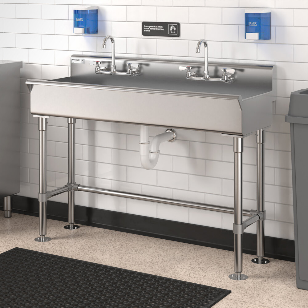 A stainless steel double hand sink with two faucets, mounted against a white tiled wall with blue glove dispensers above it.