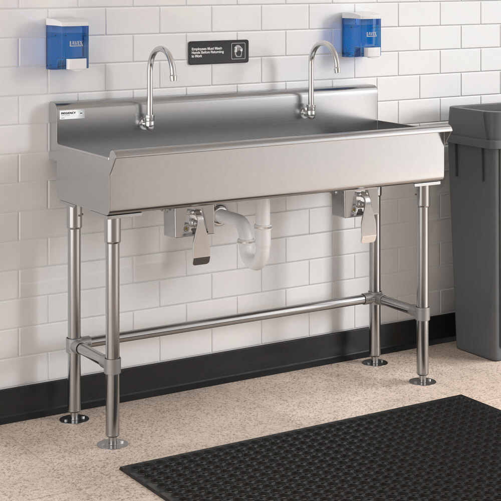 A stainless steel double faucet hand sink with foot pedal controls, mounted against a white tiled wall with soap dispensers above and a trash can nearby.