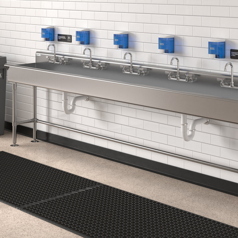 A long stainless steel hand sink with five faucets, mounted against a white tiled wall with blue soap dispensers above and black floor mats below.