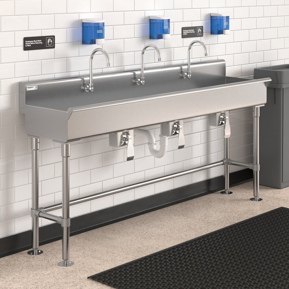 A large stainless steel hand sink with three knee-operated faucets, mounted against a tiled wall with three soap dispensers above it.