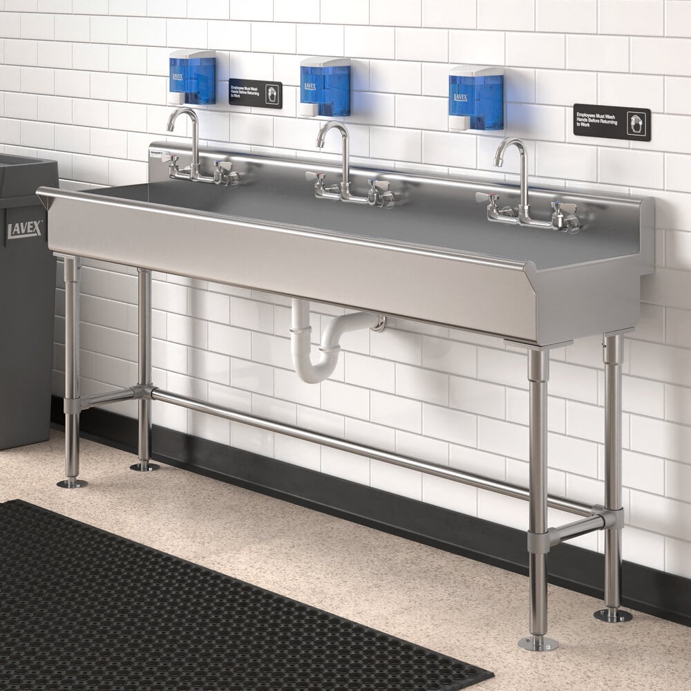 A large stainless steel hand sink with three faucets, mounted against a white tiled wall with three blue soap dispensers above it.