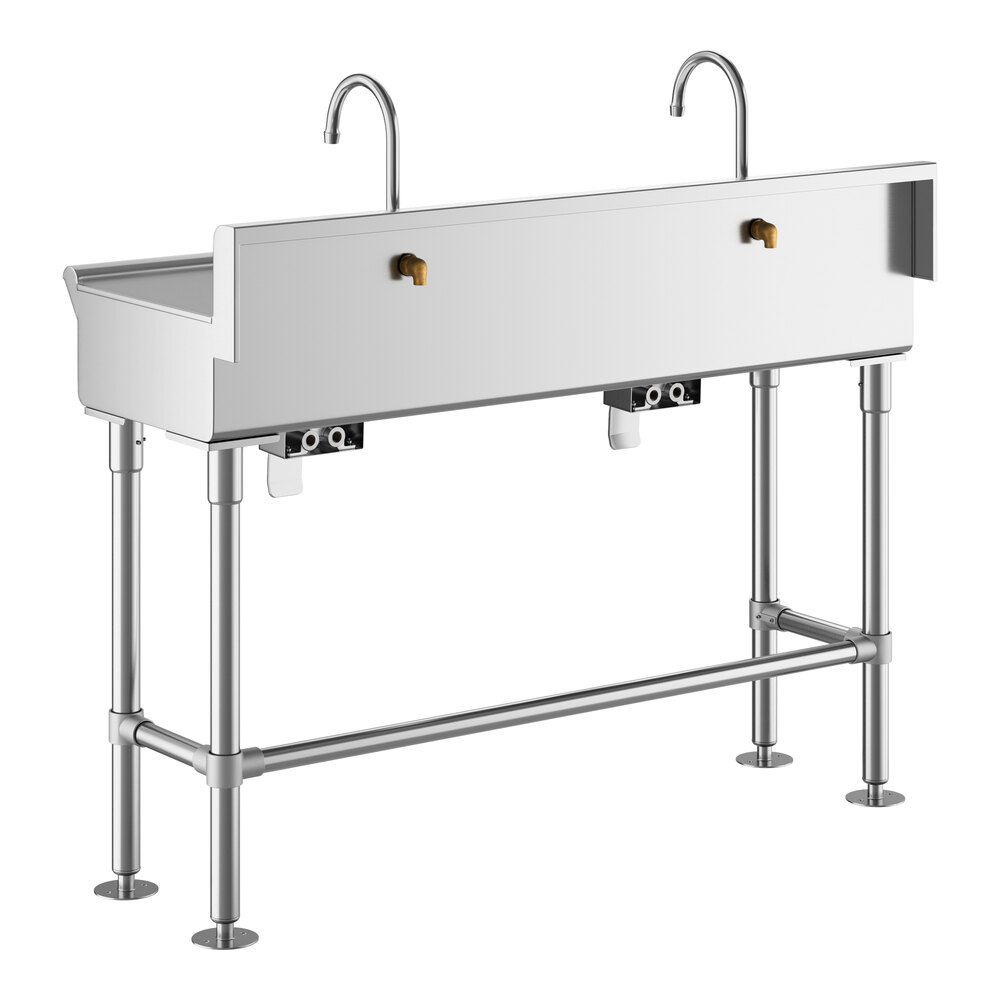 A stainless steel double-station hand sink with two gooseneck faucets and knee-operated controls, supported by tubular metal legs.