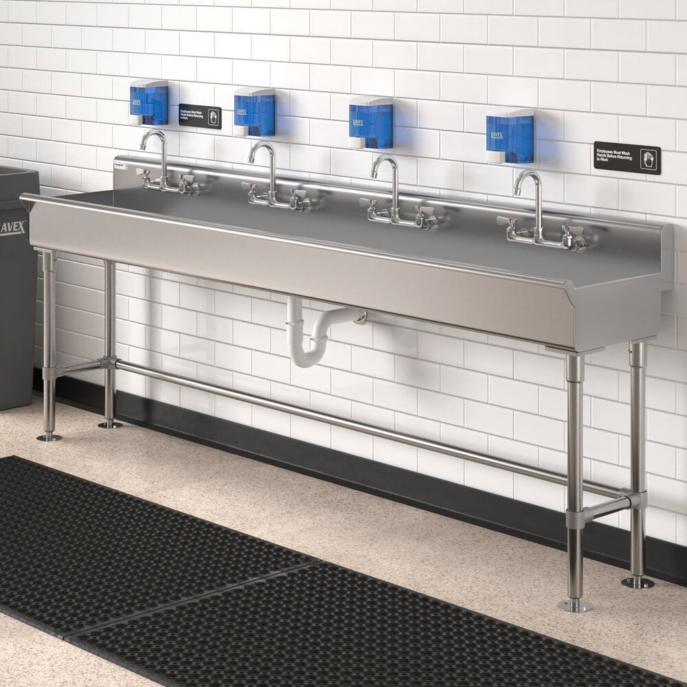 A long stainless steel hand sink with four faucets, mounted against a white tiled wall with blue soap dispensers above each faucet.