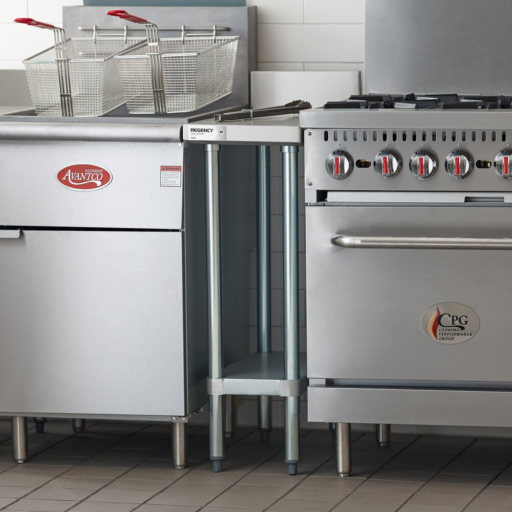 A small stainless steel filler table with an undershelf is positioned between a commercial fryer and a commercial range in a kitchen setting.