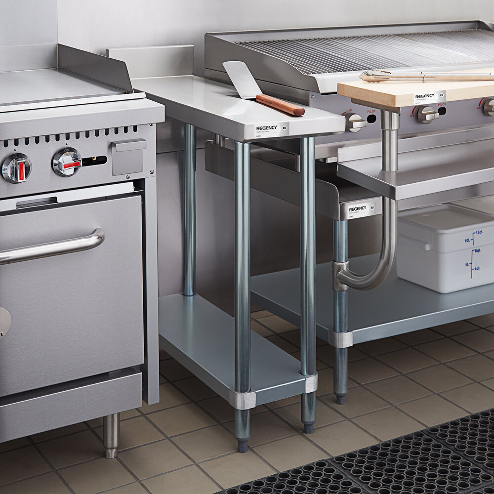 A stainless steel filler table with an undershelf is positioned between kitchen appliances in a commercial kitchen setting.