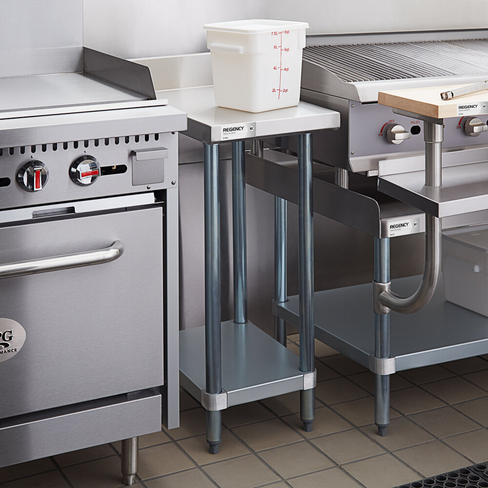 A small stainless steel table with an undershelf is positioned between kitchen appliances, with a white plastic container on top.