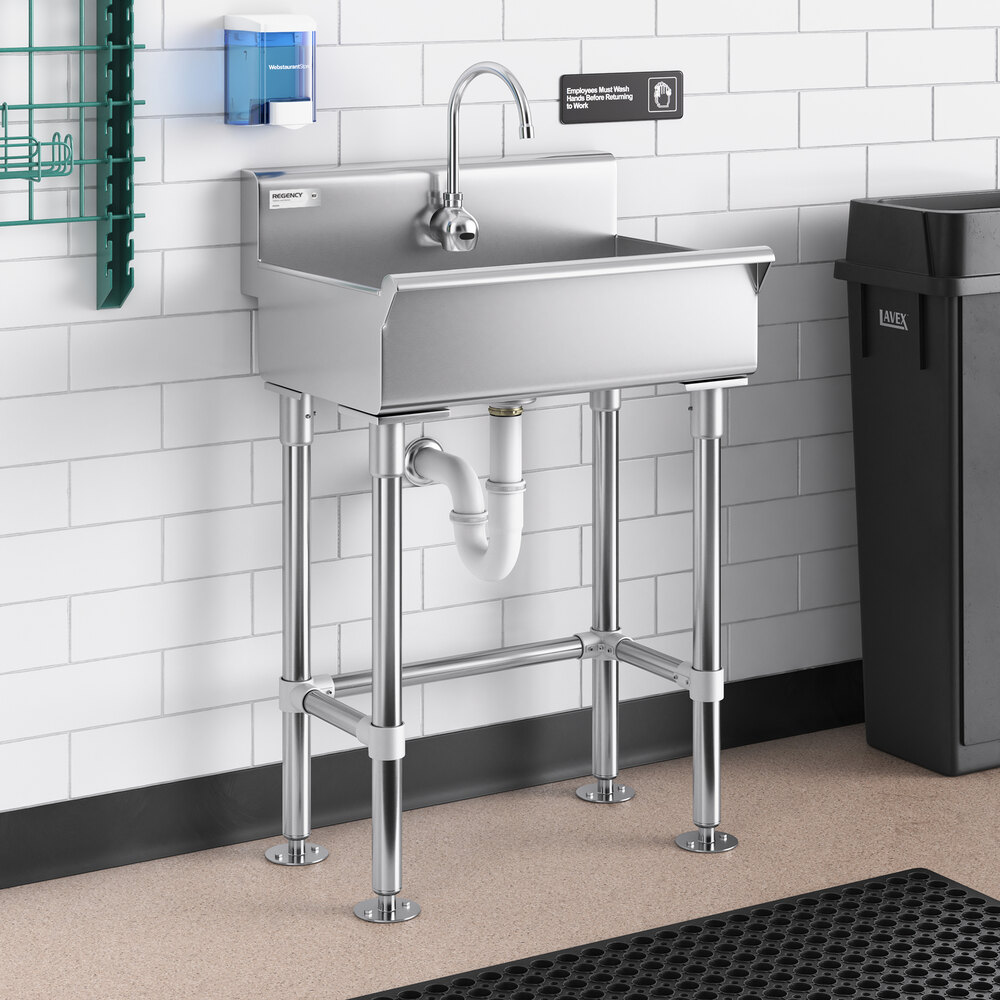 A stainless steel hand sink with a gooseneck faucet, mounted against a white tiled wall with exposed plumbing underneath.