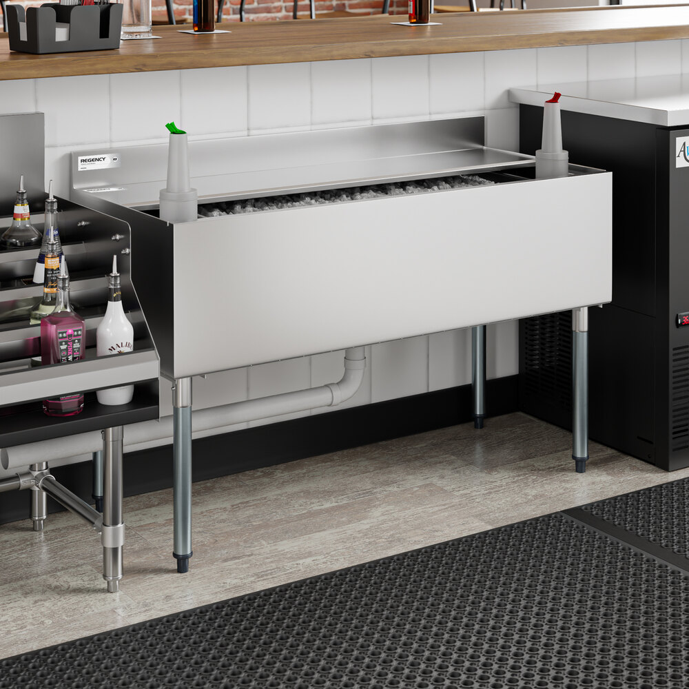 A stainless steel underbar ice bin with bottle holders and a sliding lid, partially filled with ice and positioned beneath a countertop in a bar setting.