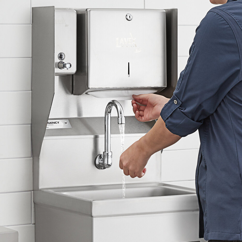 A person is washing their hands under a wall-mounted, hands-free faucet with a stainless steel sink and paper towel dispenser above.