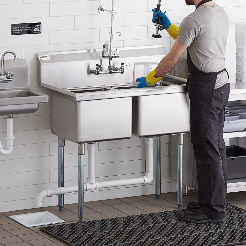 A person wearing gloves is washing items at a stainless steel two-compartment sink with metal legs in a commercial kitchen setting.