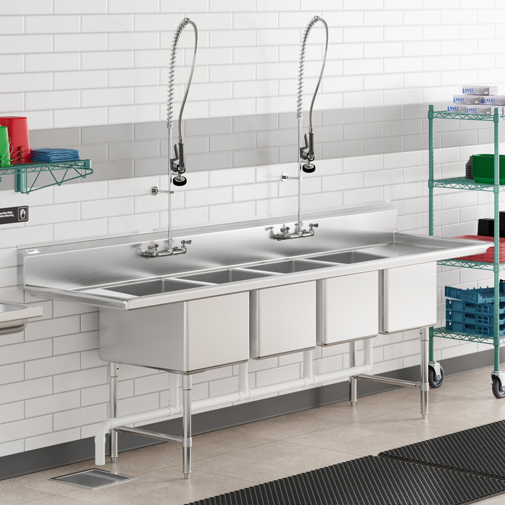 A large stainless steel commercial sink with four deep compartments, two tall pre-rinse faucets, and drainboards on both ends, set against a tiled wall in a kitchen environment.
