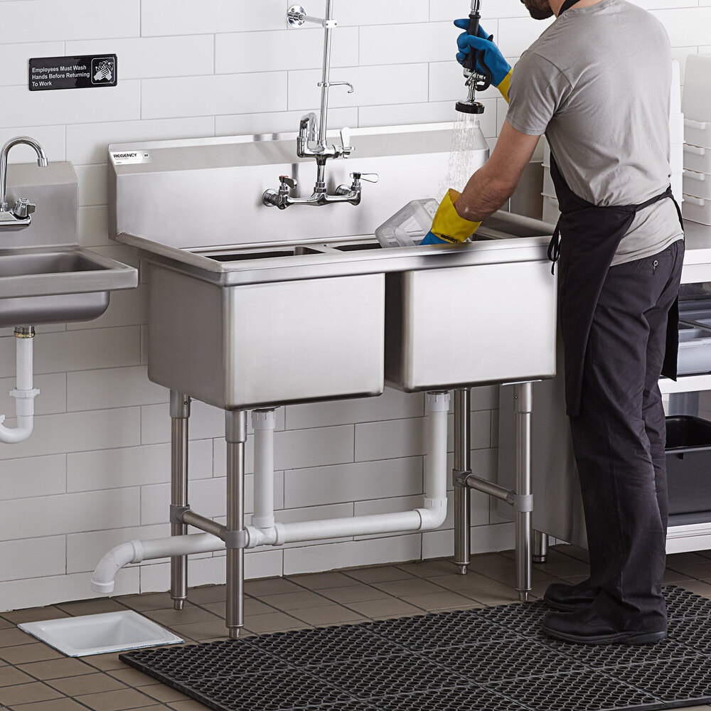A person wearing gloves and an apron is washing dishes at a stainless steel two-compartment commercial sink with visible plumbing and a sprayer attachment.