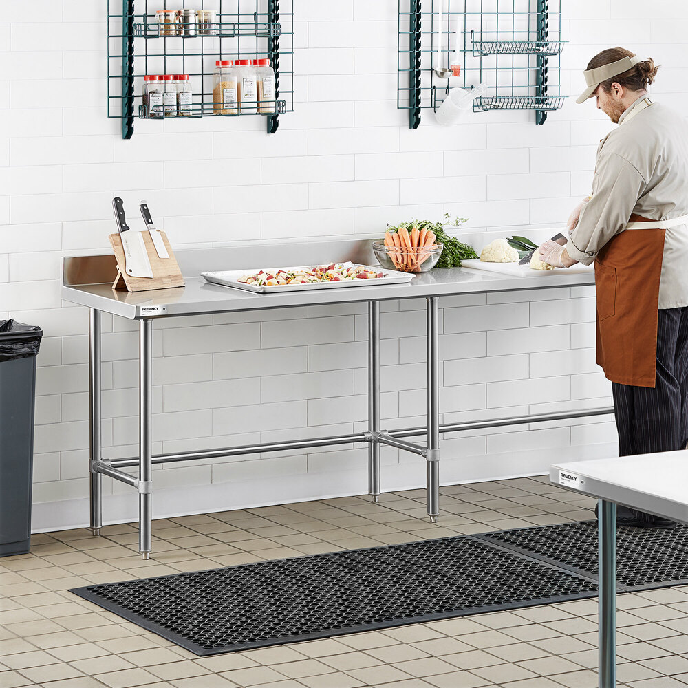 A stainless steel work table with an open base and a 4-inch backsplash, situated in a commercial kitchen setting with a person preparing vegetables.