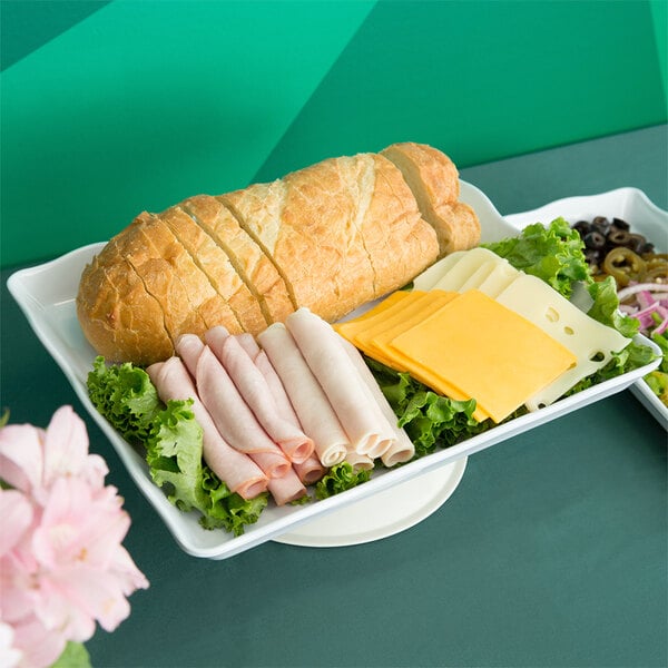 A white Osslo scalloped melamine rectangular display tray with sandwiches, cheese, and other food on a table.