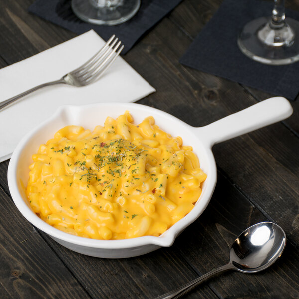 A bowl of macaroni and cheese in a Tablecraft white cast aluminum fry pan with a fork and spoon on a table.