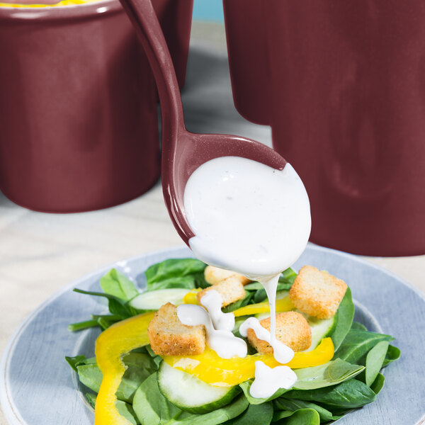 A Tablecraft maroon speckled long ladle pouring dressing over a salad.