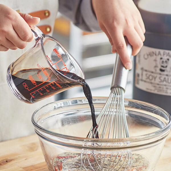 A person pouring brown soy sauce from a 1 gallon container into a bowl and using a whisk to mix it.