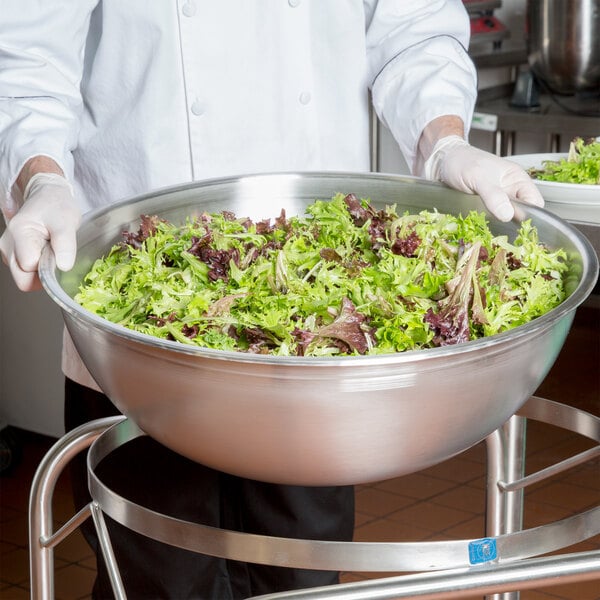 A person in a white shirt and gloves holding a Vollrath stainless steel mixing bowl filled with green salad.