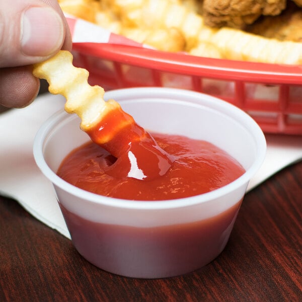 A hand holding a french fry with ketchup dipping into a Solo translucent polystyrene portion cup.
