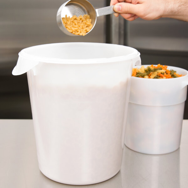 A hand pouring pasta from a measuring spoon into a white Rubbermaid food storage container with a plastic lid.